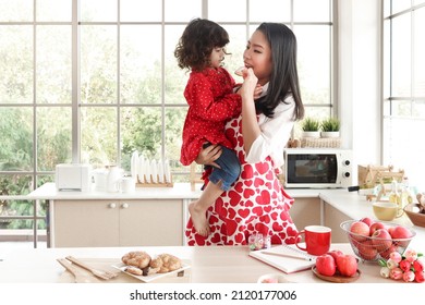 Happy Smiling Beautiful Young Asian Mom Wearing Cute Red Heart Apron Holding Little Daughter In Lovely Kitchen, Mother Feeding Her Kid With Cookie, Girl Eating Sweet From Mother Hand. 