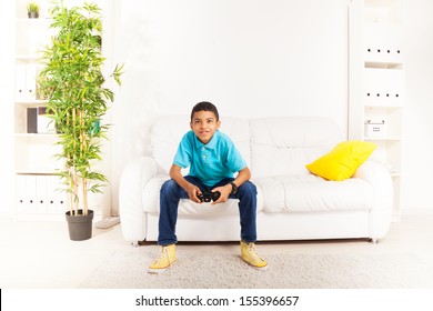 Happy Smiling Black Boy Playing Video Games Holding Game Controller Sitting On The White Sofa In Living Room