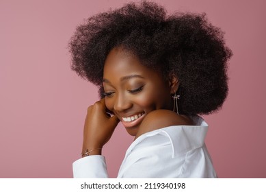Happy smiling Black woman, model wearing elegant jewelry: earrings and bracelet, white shirt, posing in studio, on pink background. Close up portrait. Copy, empty space for text