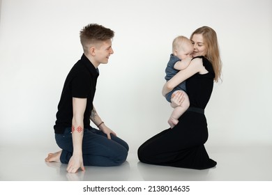 Happy Smiling Blond Family Sitting On Knee Together In Studio,holding Child In Arms. Shy Toddler Sitting Near Mum Breast