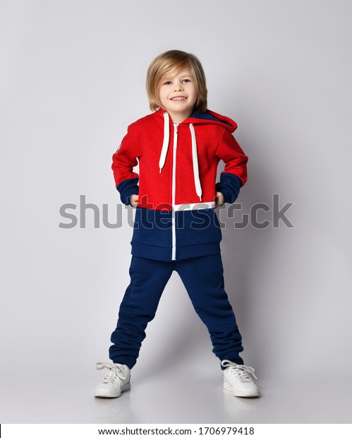 Happy smiling blond kid boy in red and blue\
sportwear and white sneakers stands with his legs wide apart and\
holding hands at his\
waist