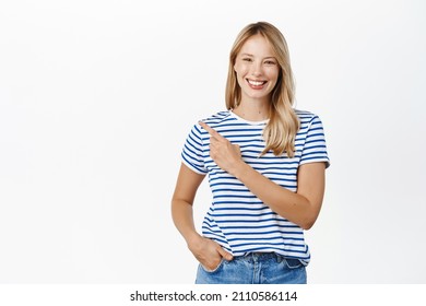 Happy smiling blond woman pointing finger left, showing announcement or banner, announcing event, standing in tshirt over white background