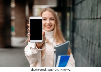 Happy Smiling Blonde Student Girl Showing Screen Of A Mobile Phone Against The Background Of The College. Mockup For Design