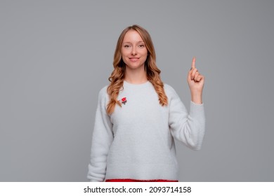 Happy smiling blonde woman pointing finger up, scolding or telling off someone, says: Attention please, wears white sweater, gray background