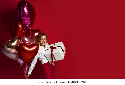 Happy smiling blonde woman in white blouse and red skirt with big gift present box and heart shaped balloons for Saint Valentine Day walks towards copy space on red background. Happy hollidays concept