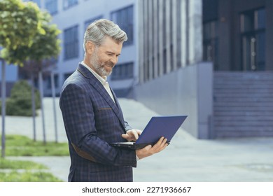 Happy and smiling boss using laptop while standing outside modern office building, mature businessman in business suit, senior investor satisfied with work result. - Powered by Shutterstock