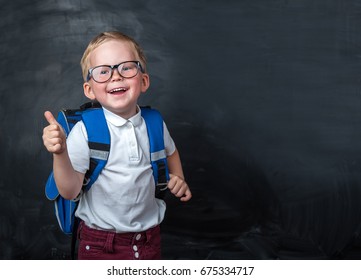 Happy smiling boy in glasses with thumb up is going to school for the first time. Child with school bag and book. Kid indoors of the class room with blackboard on a background. Back to school. - Shutterstock ID 675334717