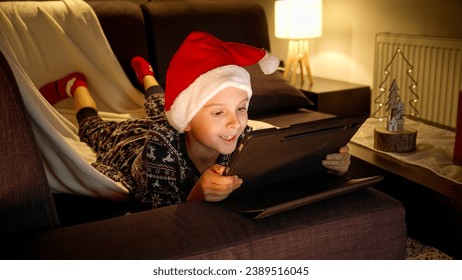 Happy smiling boy in pajamas and Santa's hat lying on sofa and using tablet computer during Christmas winter holidays. Winter holidays, celebrations and party - Powered by Shutterstock