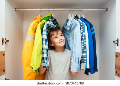 Happy Smiling Boy Searching For Clothing In A Closet. Preteen Boy Chooses Clothes In The Wardrobe Closet At Home. Kid Hiding Among Clothes In Wardrobe.