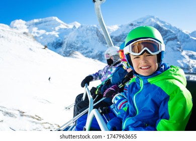 Happy Smiling Boy Sit On Chairlift Wearing Helmet And Bright Ski Outfit With Friends Over Beautiful Mountain Peak