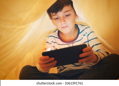 Happy Smiling Boy Sits In Bed Under A Blanket And Plays On A Smartphone In A Game In The Dark. The Child's Face Is Illuminated By A Bright Monitor