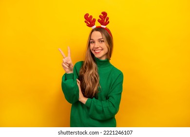 Happy smiling brunette girl showing peace gesture or v-sign with two fingers, wearing knitted green sweater with red Christmas reindeer antlers and looking at camera on a yellow background. - Powered by Shutterstock