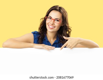 Happy smiling brunette woman in blue dress and red eye glasses, standing behind, peeping from blank banner or mock up signboard, showing pointing copy space, on yellow color background. Ophthalmology.
