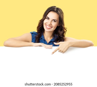 Happy smiling brunette woman in blue dress, showing, pointing, standing behind, peeping from blank banner mock up white billboard sign board signboard with ad copy space, over yellow color background.