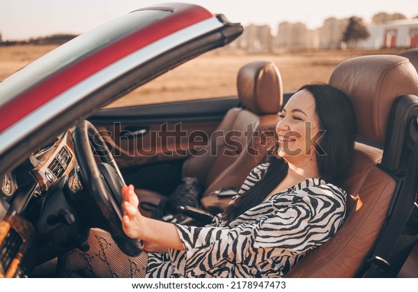 Happy smiling brunette woman driver sitting in\
new red cabriolet car on beach coast, smiling looking at camera\
enjoying journey. Driving courses and life insurance concept.\
Glamorous lifestyle