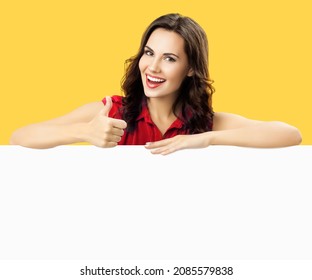 Happy smiling brunette woman in red dress cloth, showing thump up hand gesture, standing behind, peeping from blank billboard banner mock up signboard with sign copy space over yellow color background