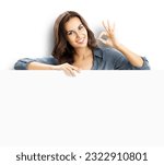 Happy smiling brunette young woman showing okay ok gesture, standing behind, peeping from blank banner or mock up signboard with copy space, isolated over white background.