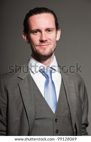 Happy smiling business man brown long hair with expressive face wearing grey suit and blue tie. Isolated on grey background.