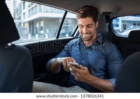 Happy smiling business man typing message on phone while sitting in a taxi. Young businessman in formal clothing using smartphone while sitting on back seat in car. Cheerful guy messaging with cell.