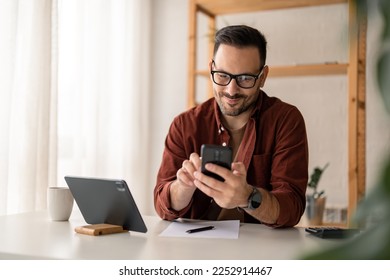 Happy smiling businessman wearing casual clothes and using modern smartphone in his home office during the day, typing, touching the screen, browsing the internet or writing text messages.