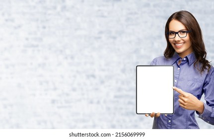 Happy Smiling Businesswoman In Eye Glasses Spectacles Showing Touchpad Tablet Pc, With White Screen Copy Space Text Area Advertising Or Slogan Sign, Over White Bricks Wall. Skype Zoom Chat Interview.