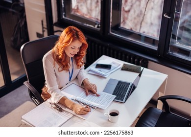 Happy smiling businesswoman in formal wear sitting at desk in modern office in front of laptop and doing paperwork - Shutterstock ID 2256175863