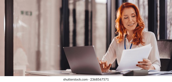 Happy smiling businesswoman in formal wear sitting at desk in modern office in front of laptop and doing paperwork - Shutterstock ID 2368567571
