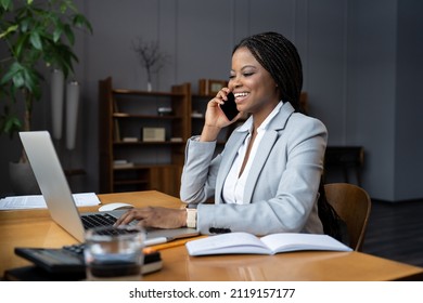 Happy Smiling Businesswoman Talk On Phone Sitting At Workplace. Joyful Young African American Lady Business Owner In Formal Wear Calling Friend, Family Or Satisfied Client Working On Laptop In Office