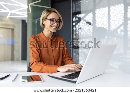 Happy and smiling businesswoman typing on laptop, office worker with glasses happy with achievement results, at work inside office building