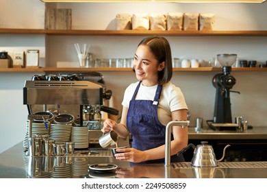 Happy smiling cafe owner, girl barista in apron, making cappuccino, latte art with steamed milk, standing behind counter. - Powered by Shutterstock