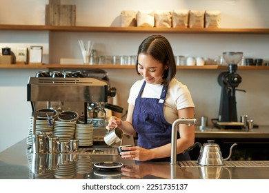 Happy smiling cafe owner, girl barista in apron, making cappuccino, latte art with steamed milk, standing behind counter. - Powered by Shutterstock