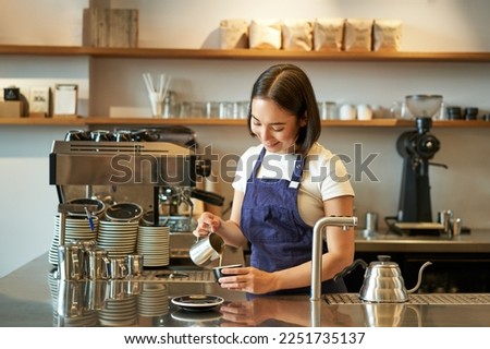 Happy smiling cafe owner, girl barista in apron, making cappuccino, latte art with steamed milk, standing behind counter.