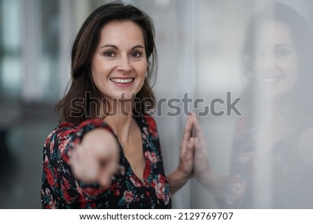 happy smiling casual business woman pointing finger at camera to address you directly for motivation surrounded by modern white indoor walls
