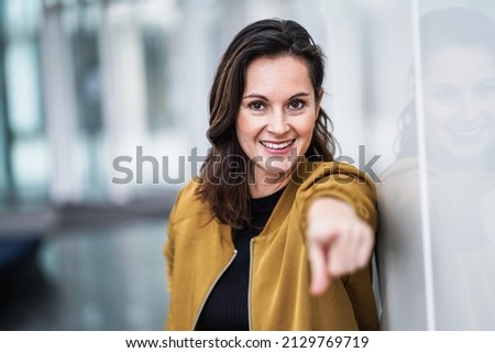 happy smiling casual business woman pointing finger at camera to address you directly for motivation surrounded by modern white indoor walls
