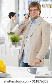 Happy Smiling Caucasian Architect At Bright Office Talking On Mobile Phone, Pointing At Table, Standing. Suit No Tie, Looking At Camera.