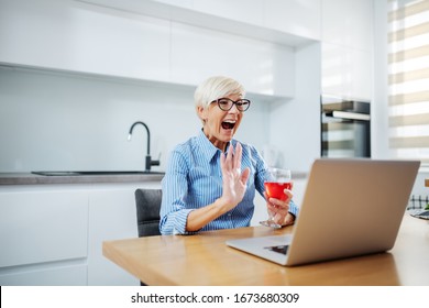 Happy Smiling Caucasian Blonde Senior Woman Sitting At Dining Table, Drinking Red Wine, Having Video Call Over At Laptop And Waving.