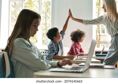 Happy smiling Caucasian female teacher giving high five to asian schoolboy standing in classroom with group of schoolchildren. Teamwork successful lesson. Technologies for education concept. - Powered by Shutterstock