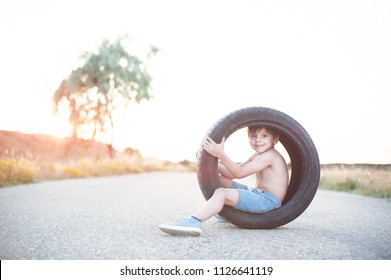 Happy Smiling Caucasian Little Kid In Shorts Sitting Inside Car Tire On Empty Asphalt Road