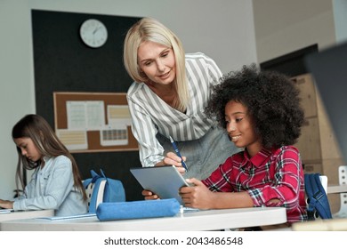 Happy Smiling Caucasian Teacher Explaining Task To African American Schoolgirl Using Tablet Device Sitting In Classroom With Group Of Schoolchildren. Modern Technologies For Education Concept.