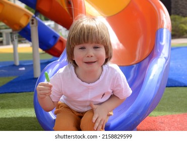 Happy Smiling Child 3 Years Old, Blond. A Cute Boy Laughs On The Playground And Holds A Green Lolipop In His Hand. Concept: Good Mood, Family Vacation