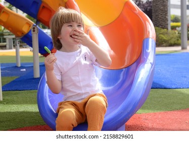 Happy Smiling Child 3 Years Old, Blond. A Cute Boy Laughs On The Playground And Holds A Green Lolipop In His Hand. Concept: Good Mood, Family Vacation