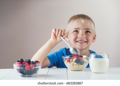 Happy, smiling child eating oatmeal with berries. The concept of a healthy breakfast for children, porridge with raspberries and blackberries, a glass of yogurt on white wooden table.