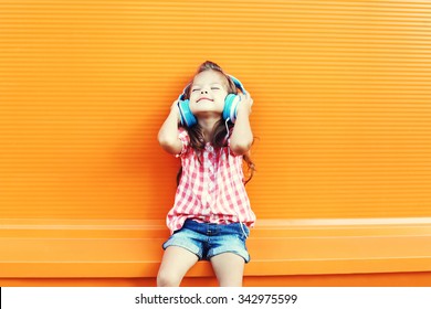 Happy Smiling Child Enjoys Listens To Music In Headphones Over Orange Background