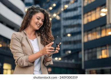 Happy smiling confident businesswoman standing in front of the modern workplace in big city using smartphone for texting, cellphone apps for work outside.