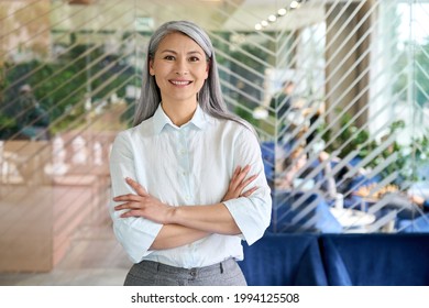 Happy Smiling Confident Middle Aged Asian Older Senior Female Leader Businesswoman Standing In Modern Office Workplace Looking At Camera Arms Crossed. Business Successful Executive Concept. Portrait.