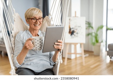 Happy smiling confident senior blonde woman sitting relaxed in swing drinking coffee and surfing on digital tablet in living room in her apartment. - Powered by Shutterstock