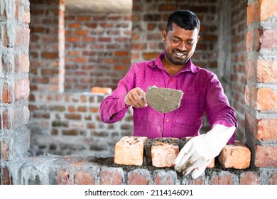 Happy smiling construction worker constructing or building wall by placing bricks and cement - concept of hard working, manual labour and daily wagers lifestyle