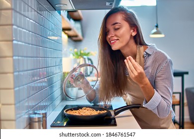 Happy Smiling Cooking Woman Housewife Preparing Food In A Frying Pan On The Stove For Delicious Dinner And Enjoying Of Smell Of Cooked Dish At Modern Loft Style Kitchen  