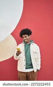 Happy smiling cool gen z young African American ethnic stylish hipster guy model standing at red city urban wall outdoors using cell phone mobile device, looking at camera holding cellphone, vertical. - Shutterstock ID 2272222455