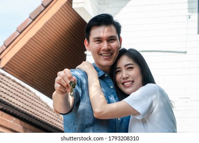 Happy Smiling Couple In Front Of The Home And Holding Keys To New Home On Moving In Day, And Start A New Life Family. Concept Of Homeowner And Relocation